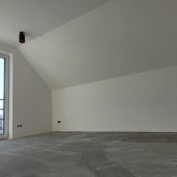 Gfk Design Grzegorz Kowalski - Poddasze z oknem balkonowym, świeżo pomalowane na biało, z widocznymi gniazdkami elektrycznymi i szarą posadzką. W tle widoczny budynek.