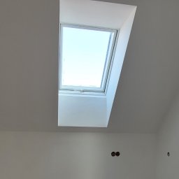 Gfk Design Grzegorz Kowalski - Białe poddasze z oknem dachowym, widoczne instalacje elektryczne i hydrauliczne, surowa podłoga przed wykończeniem. Ściany świeżo pomalowane.