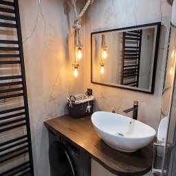 Gfk Design Grzegorz Kowalski - Nowoczesna łazienka z umywalką nablatową, blatem z ciemnego drewna i lampą ze sznura. W tle marmurowe płytki i czarny grzejnik drabinkowy. Widoczna pralka.