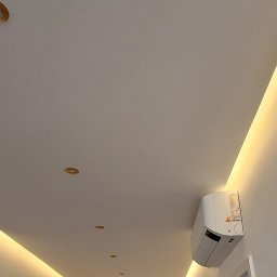 Gfk Design Grzegorz Kowalski - Biały sufit z oświetleniem LED wzdłuż krawędzi, z klimatyzatorem na ścianie. Widoczny fragment korytarza z drzwiami i podłogą z paneli.