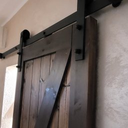 Gfk Design Grzegorz Kowalski - Drewniane drzwi przesuwne w stylu rustykalnym z czarnymi okuciami, zamontowane na ścianie o fakturze baranka. Widok z dołu, na pierwszym planie fragment ściany.