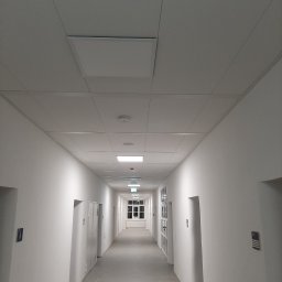 Gfk Design Grzegorz Kowalski - Długi, biały korytarz z równomiernie rozmieszczonymi drzwiami i podwieszanym sufitem z panelami. Widoczne oświetlenie sufitowe i okno na końcu korytarza.