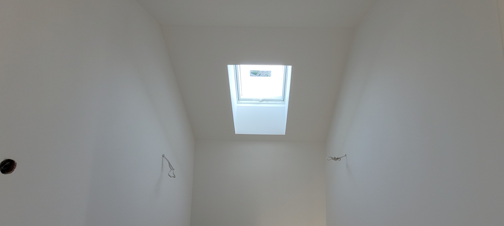 Białe, świeżo pomalowane poddasze z oknem dachowym. Widoczne wystające kable i otwór na gniazdko, sugerujące prace wykończeniowe.