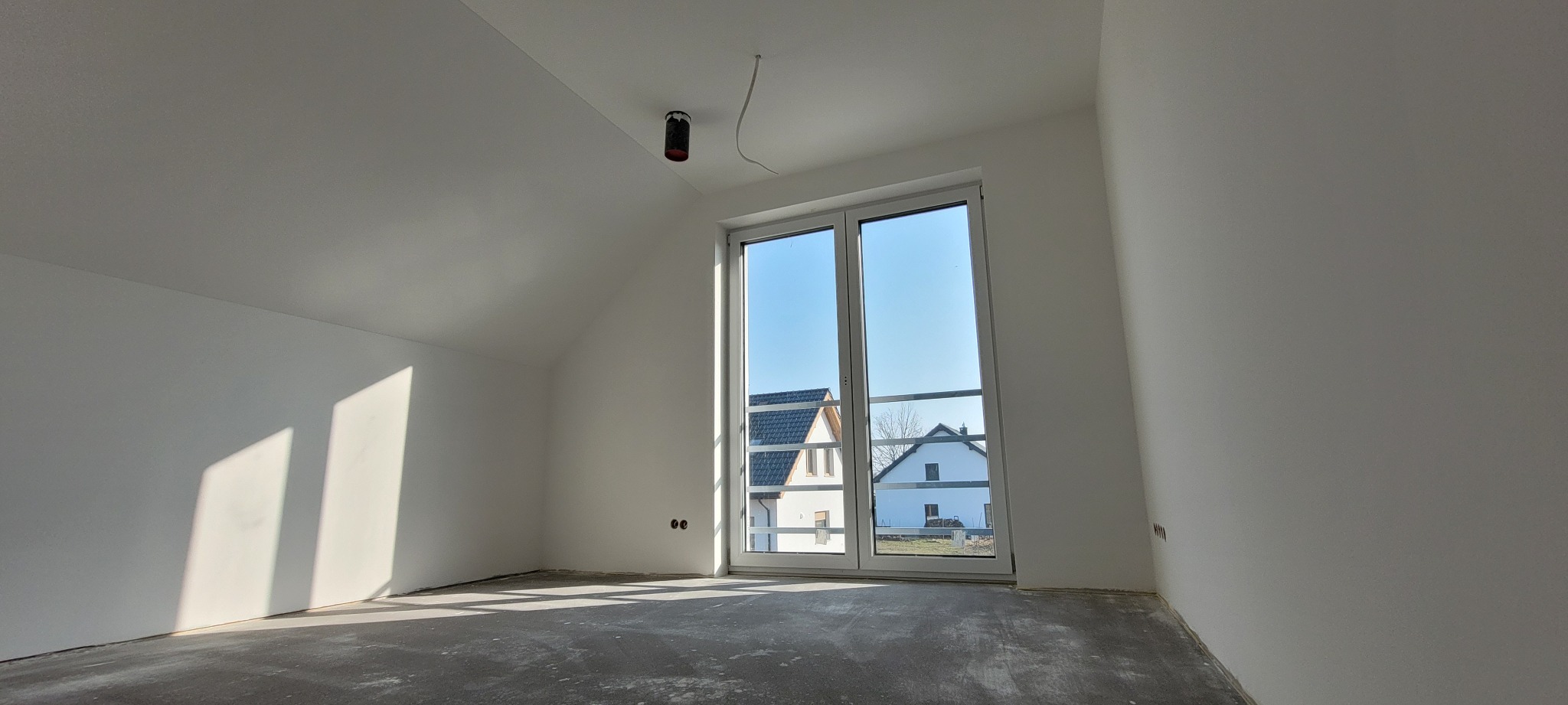 Jasne, świeżo pomalowane poddasze z oknem balkonowym i widokiem na domy. Ściany białe, podłoga szara, surowa. Widoczne przygotowania do wykończenia wnętrza.