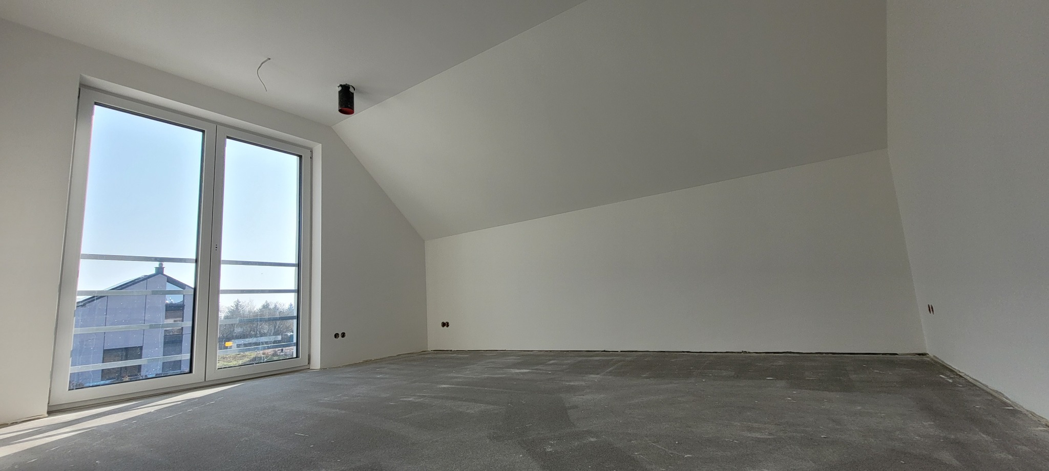 Poddasze z oknem balkonowym, świeżo pomalowane na biało, z widocznymi gniazdkami elektrycznymi i szarą posadzką. W tle widoczny budynek.