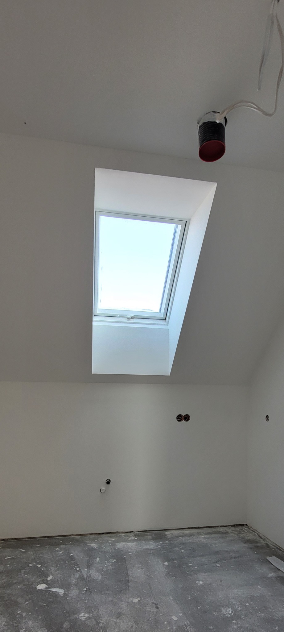 Białe poddasze z oknem dachowym, widoczne instalacje elektryczne i hydrauliczne, surowa podłoga przed wykończeniem. Ściany świeżo pomalowane.