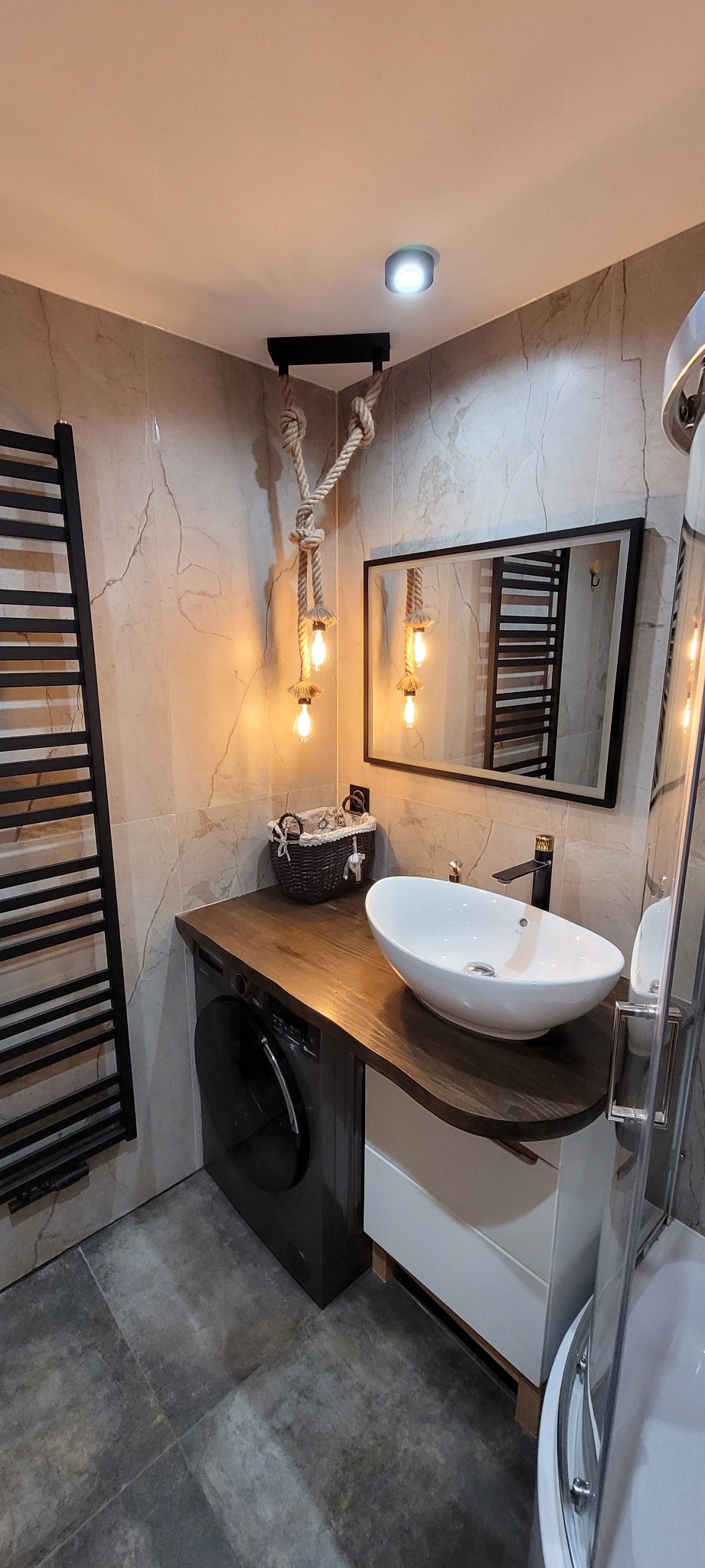 Nowoczesna łazienka z umywalką nablatową, blatem z ciemnego drewna i lampą ze sznura. W tle marmurowe płytki i czarny grzejnik drabinkowy. Widoczna pralka.