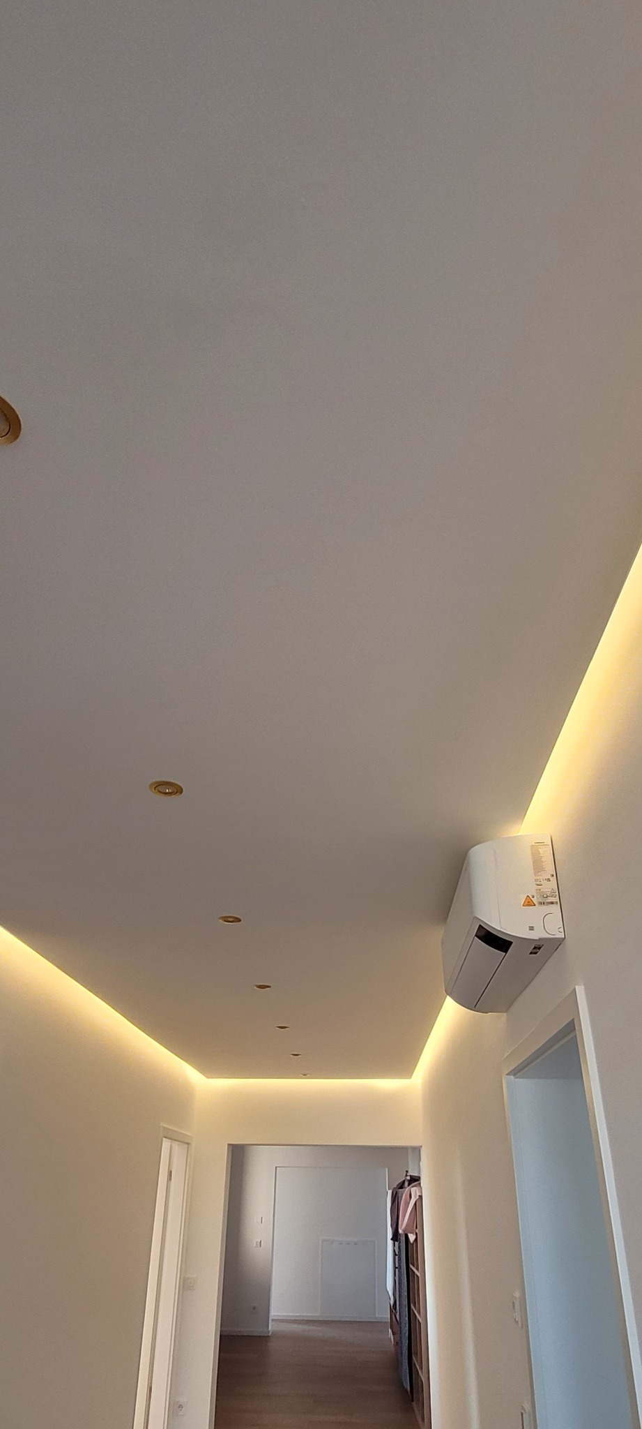 Biały sufit z oświetleniem LED wzdłuż krawędzi, z klimatyzatorem na ścianie. Widoczny fragment korytarza z drzwiami i podłogą z paneli.