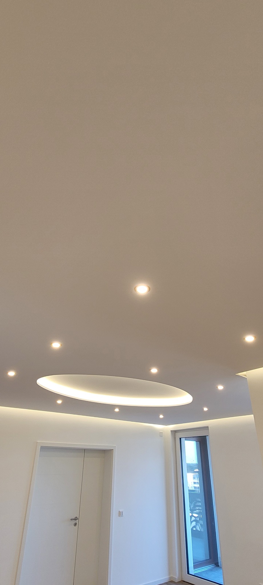 Biały sufit z owalnym podświetleniem LED i punktowymi lampami, widok na białe ściany, drzwi i okno balkonowe. Nowoczesne wykończenie wnętrza.