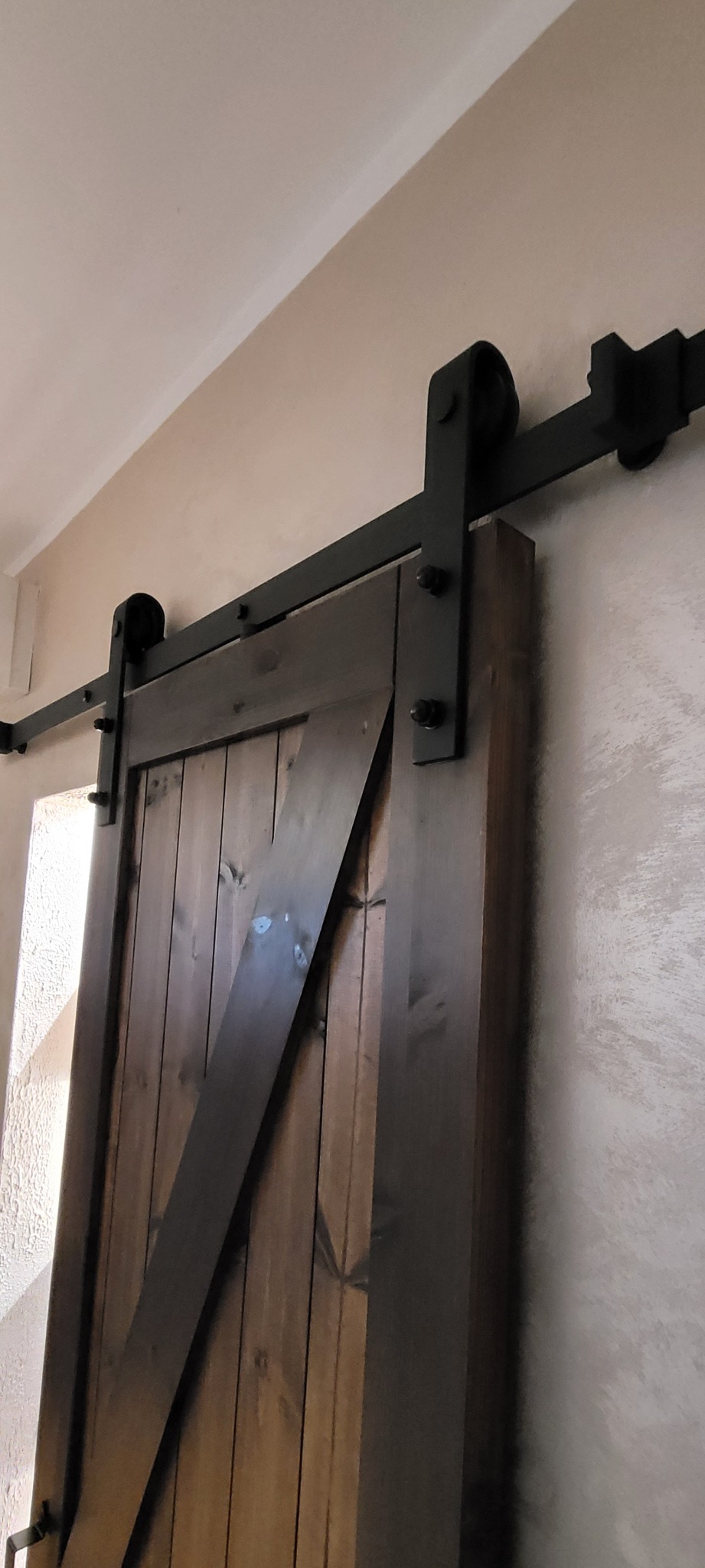 Drewniane drzwi przesuwne w stylu rustykalnym z czarnymi okuciami, zamontowane na ścianie o fakturze baranka. Widok z dołu, na pierwszym planie fragment ściany.