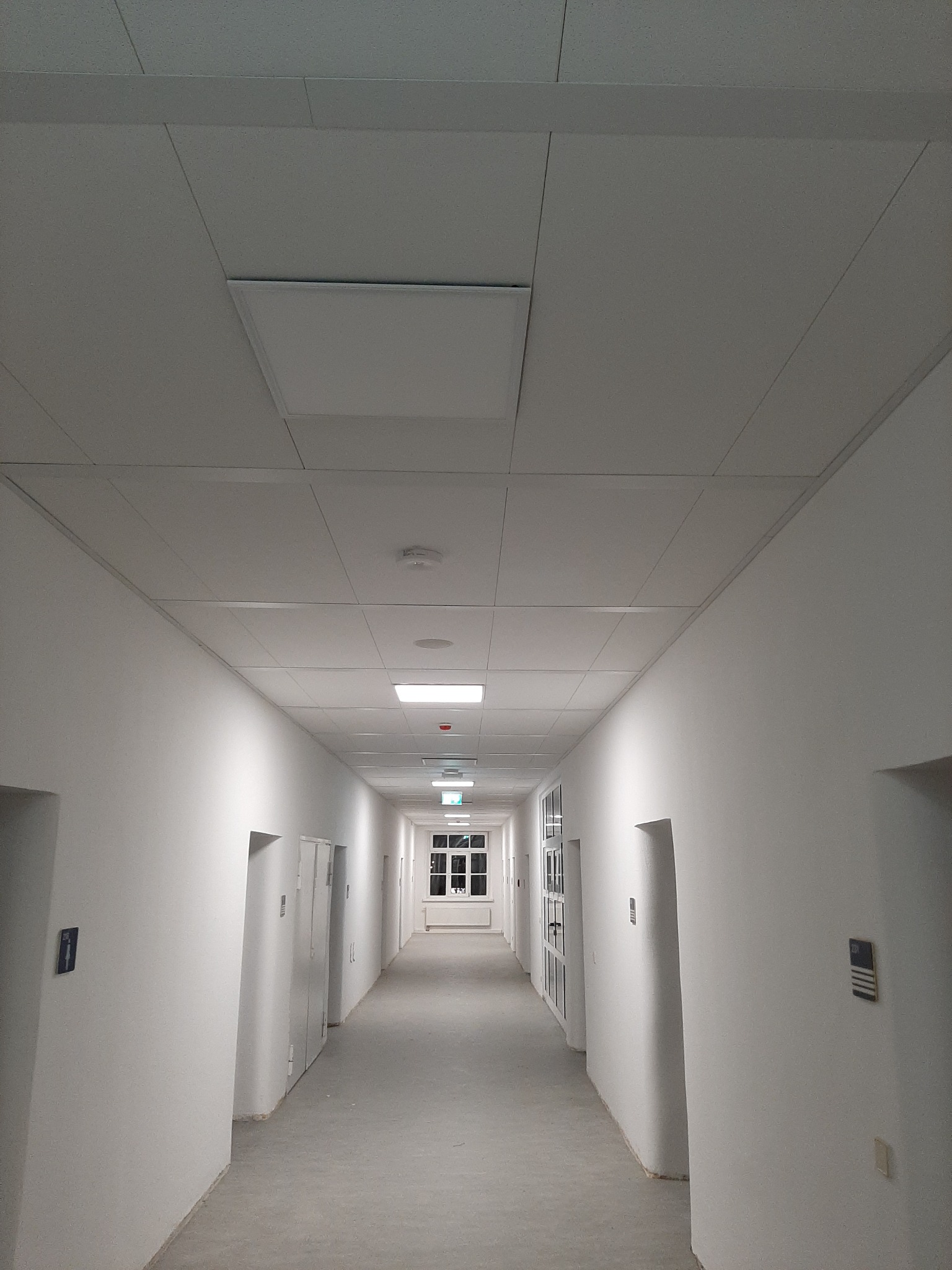 Długi, biały korytarz z równomiernie rozmieszczonymi drzwiami i podwieszanym sufitem z panelami. Widoczne oświetlenie sufitowe i okno na końcu korytarza.