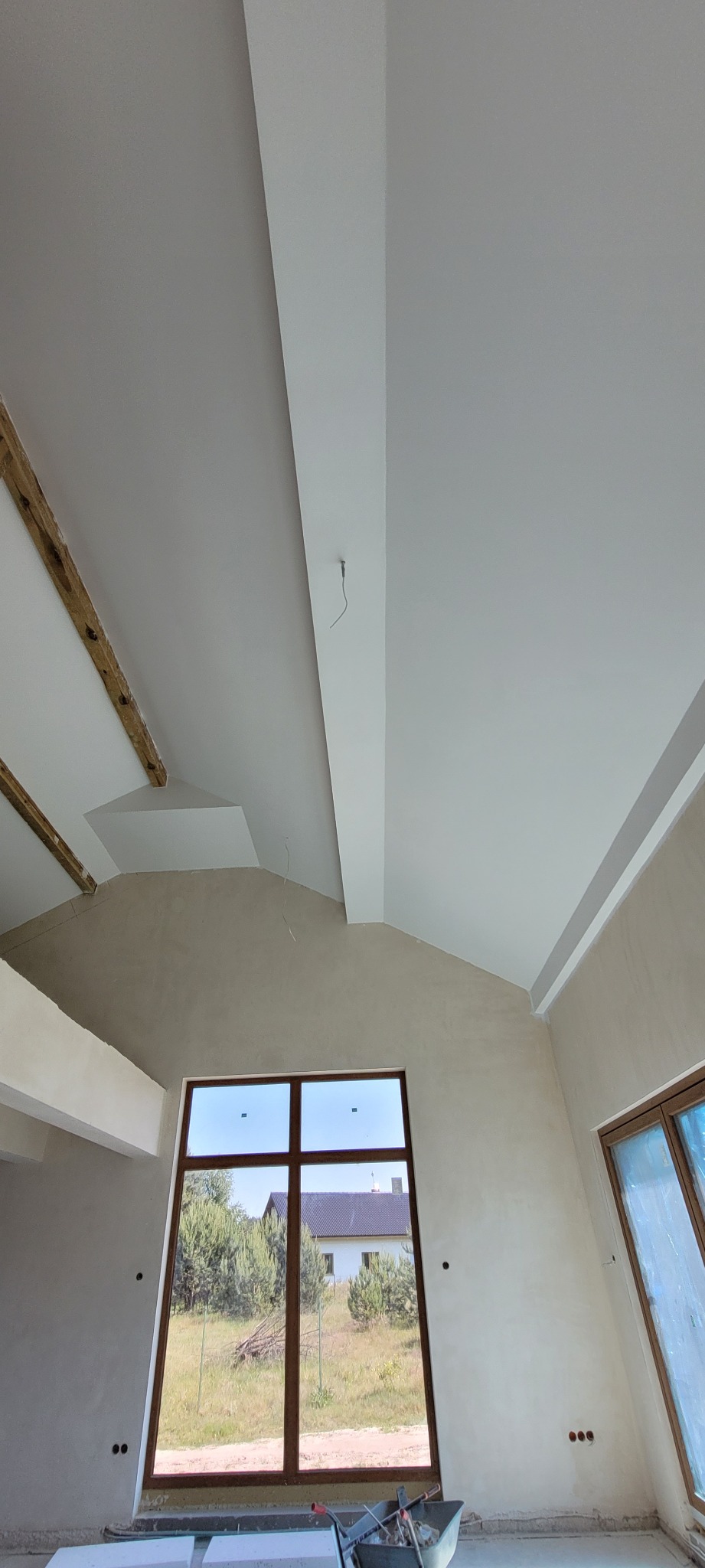 Wnętrze w trakcie wykończenia: gładkie, białe sufity z elementami drewnianymi i gipsowymi, ściany przygotowane do malowania, duże okno z widokiem na zewnątrz.