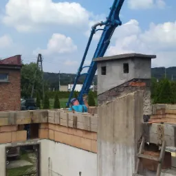 Budowa domu jednorodzinnego: murowanie ścian z bloczków ceramicznych, widoczna konstrukcja stropu z drewnianych belek i szalunków, na tle budynek z czerwonej cegły i ramię pompy do betonu.