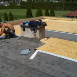 Dekarze układają papę termozgrzewalną na dachu z płyt OSB, widoczny komin i częściowo ułożona izolacja.