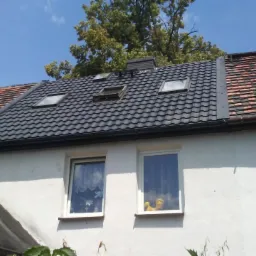 Częściowo odnowiony dach z czarną dachówką i oknami dachowymi, sąsiadujący ze starą dachówką ceramiczną na budynku z białymi ramami okiennymi.