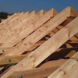 Świeżo zmontowana, jasna więźba dachowa z belek drewnianych, połączonych metalowymi śrubami, na tle błękitnego nieba i zabudowań.