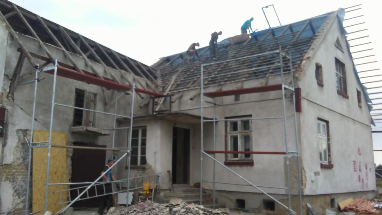 Ekipa budowlana na rusztowaniu podczas prac na dachu starego, częściowo rozebranego domu. Widoczne elementy więźby dachowej i fragmenty starego pokrycia.