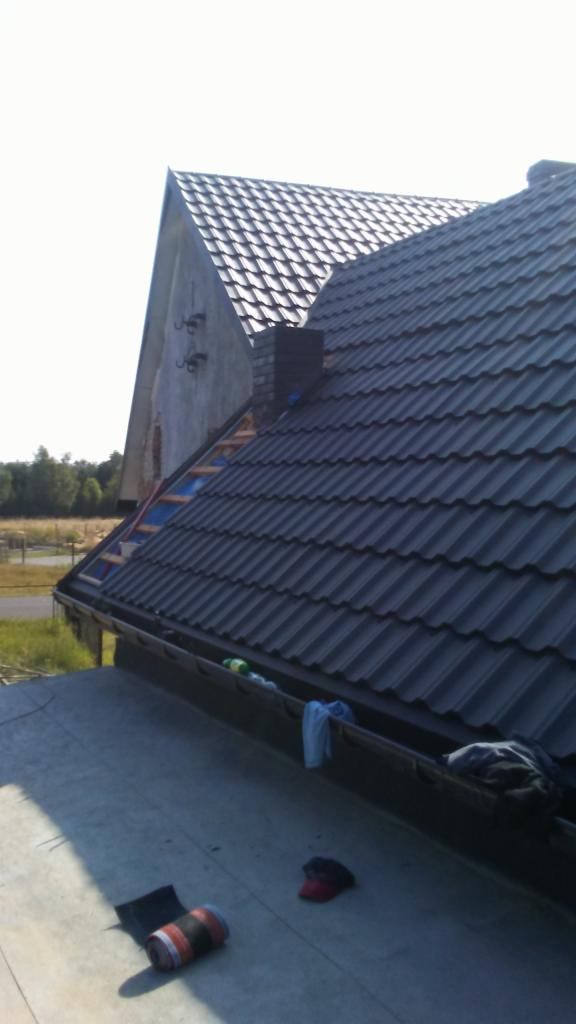 Ciemnoszary dach z dachówki ceramicznej, widok z góry na rynny z ubraniami i roboczymi akcesoriami, w tle szczyt budynku z częściowo zdjętym poszyciem i las.