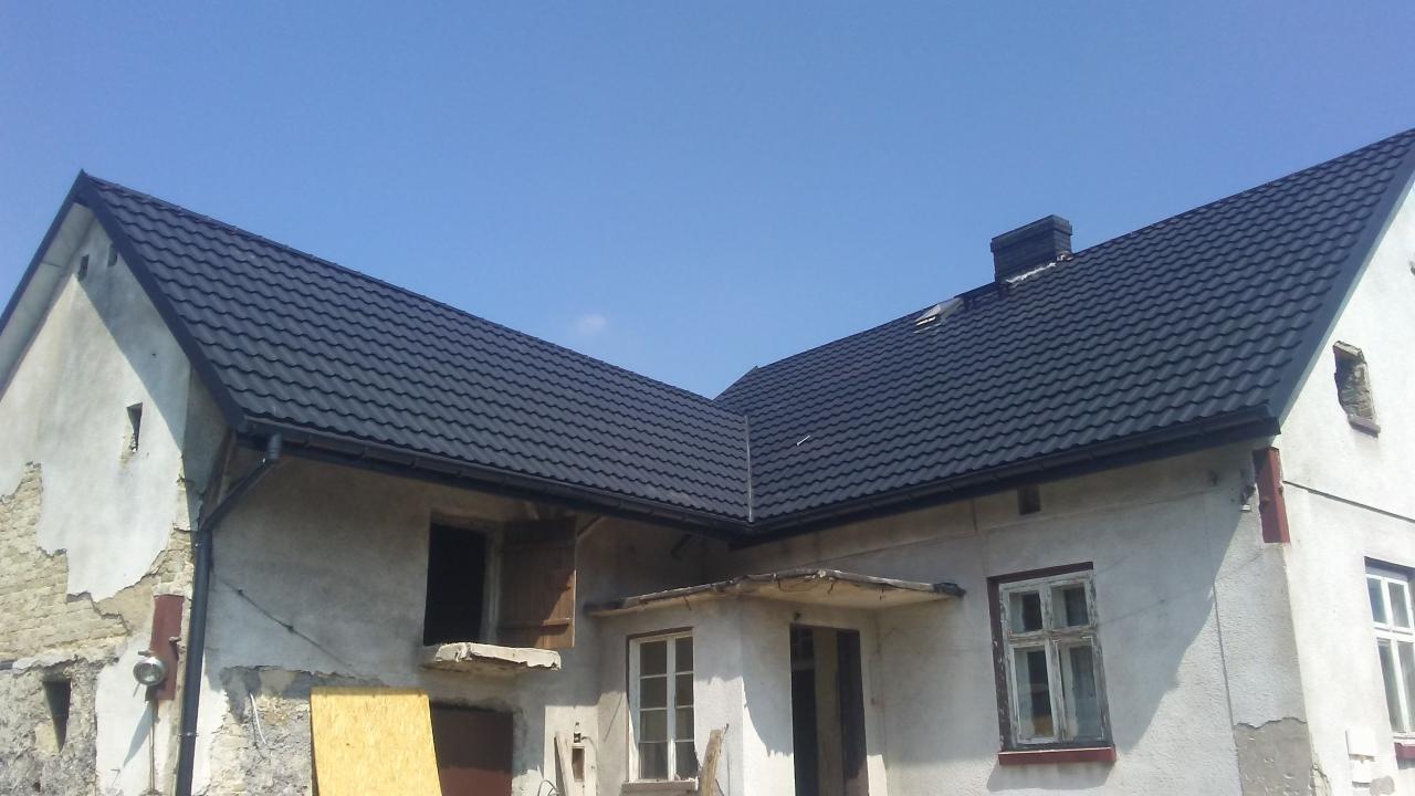 Nowy, ciemny dach z blachodachówki na starym, częściowo otynkowanym domu z odsłoniętą cegłą i niezabezpieczonymi oknami, widok z dołu.