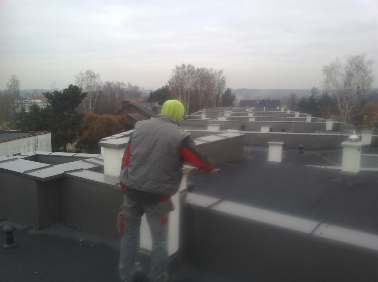 Pracownik w odblaskowej kamizelce i zielonym kapturze na płaskim dachu z licznymi kominami, wykonujący prace wykończeniowe przy obróbce blacharskiej, w tle widok na zabudowę miejską i drzewa...