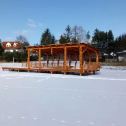 Drewniana konstrukcja altany z miejscami do siedzenia, umieszczona na zamarzniętym jeziorze pokrytym śniegiem, w tle domy i drzewa.