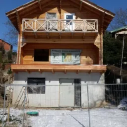 Dwupiętrowy dom z bali w trakcie budowy, widoczne elementy konstrukcyjne, niezabezpieczone otwory okienne, na elewacji baner reklamowy producenta, teren wokół domu częściowo pokryty śniegiem...