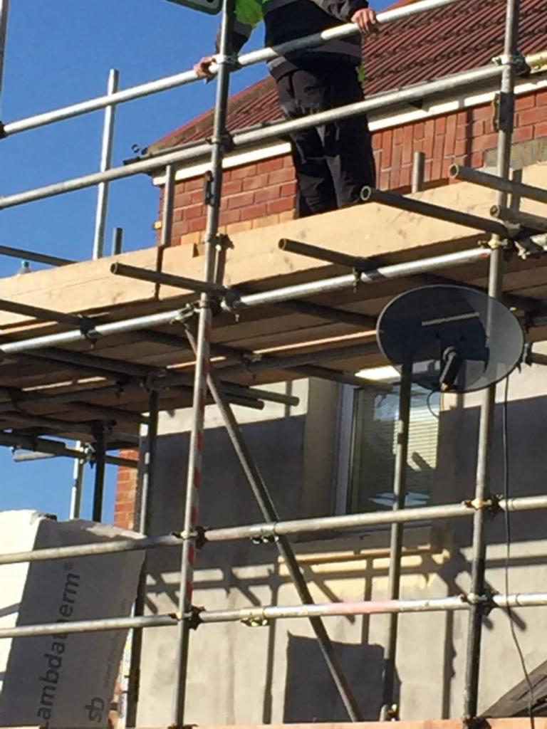 Fragment rozbudowy domu z widocznym rusztowaniem, robotnikiem w odblaskowej kamizelce na górze, ociepleniem LambdaTherm i satelitarną anteną na elewacji.
