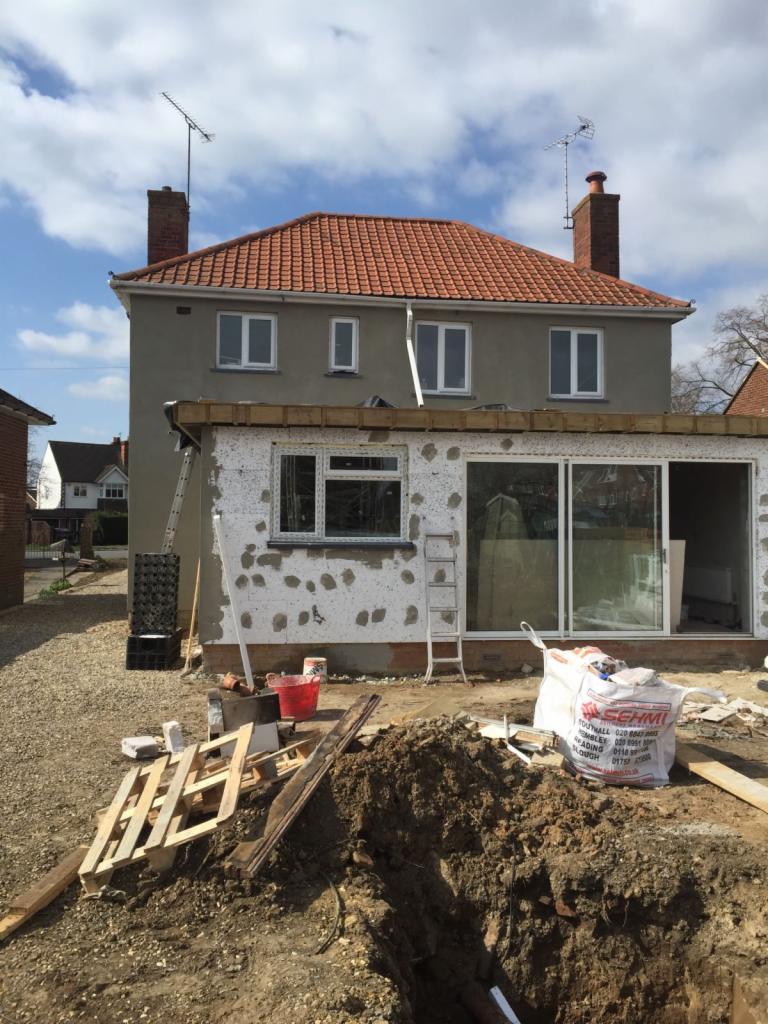 Dom z szarą elewacją i czerwoną dachówką w trakcie rozbudowy, widoczne okna PCV, rusztowania, materiały budowlane i wykop ziemny przed budynkiem.