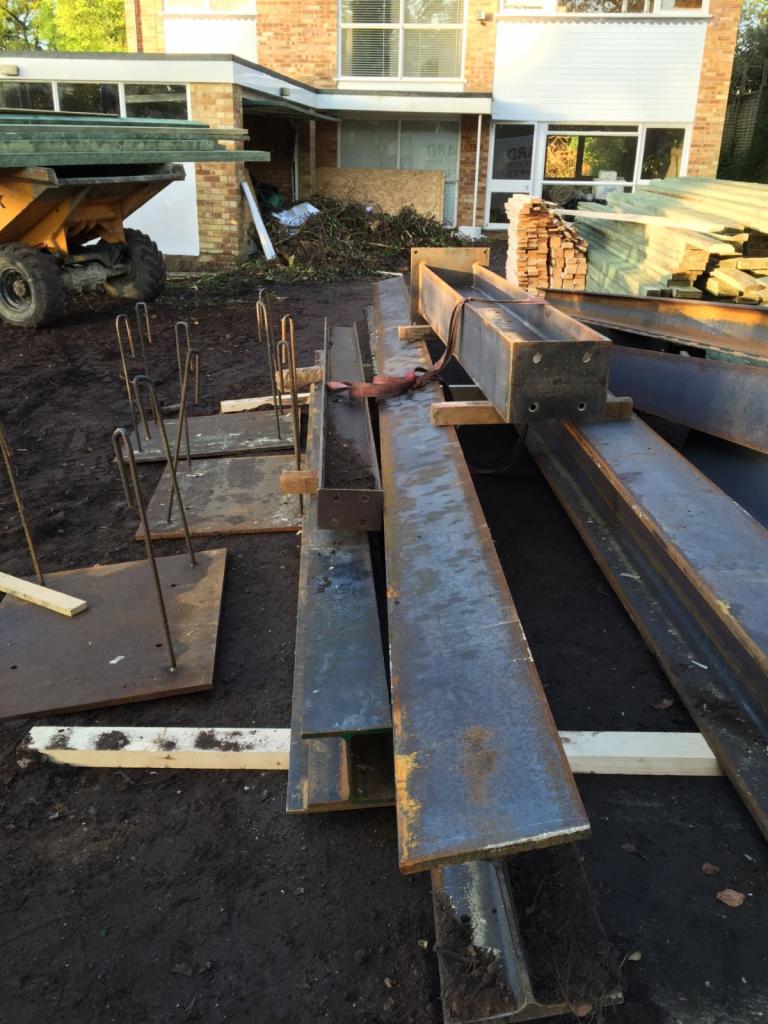 Przygotowane stalowe belki i pręty zbrojeniowe na placu budowy przed domem z cegły, żółta wywrotka w tle.