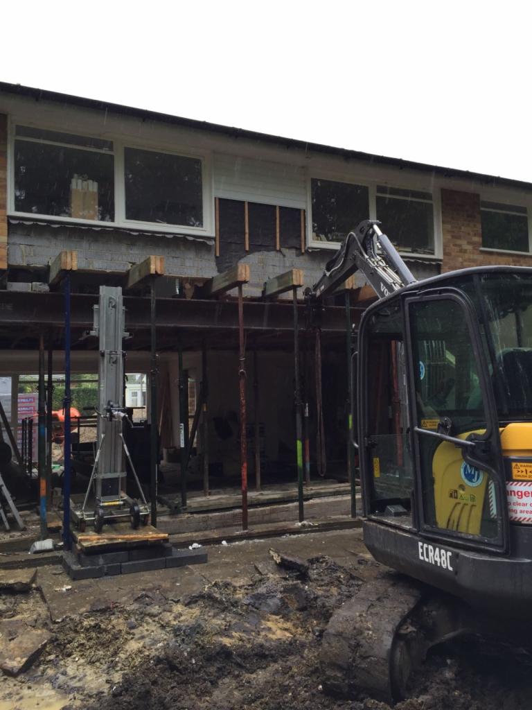 Prace przygotowawcze do rozbudowy murowanego domu z dużą ilością okien, widoczny ciężki sprzęt budowlany i podpory konstrukcyjne.