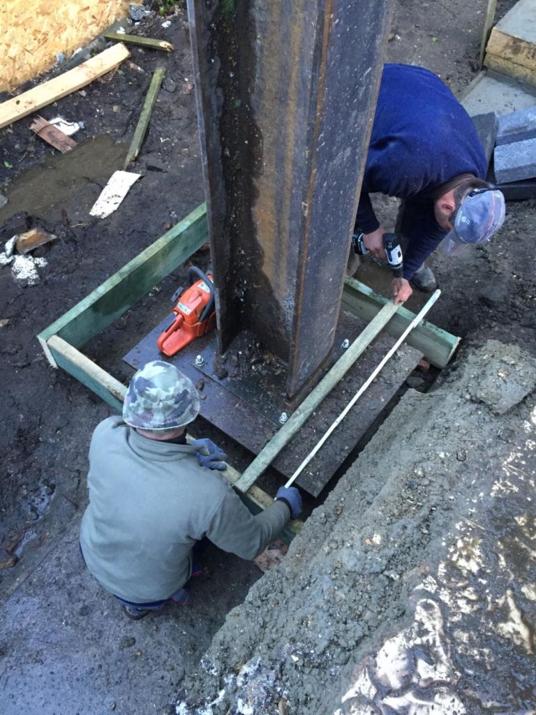 Montaż stalowej kolumny na fundamencie: dwóch pracowników mocuje drewniane szalunki, widoczna piła spalinowa, teren budowy z błotem i fragmentami betonu.