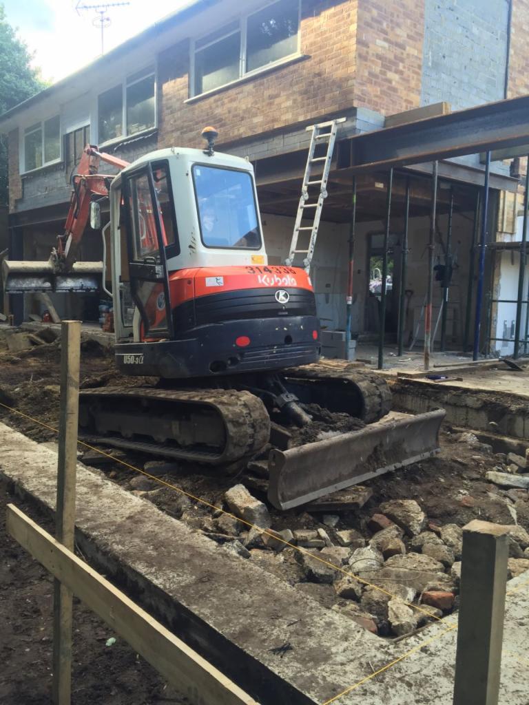 Mini koparka Kubota na placu budowy podczas prac ziemnych przy rozbudowie domu z cegły, widoczne stalowe belki i rusztowanie.