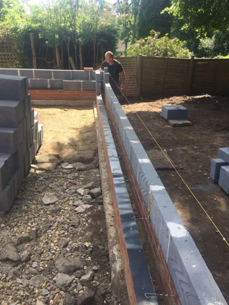 Wznoszenie muru z bloczków betonowych i cegły, widoczny pracownik z cegłą w dłoni, sznur murarski naprężony wzdłuż ściany, przygotowany fundament.