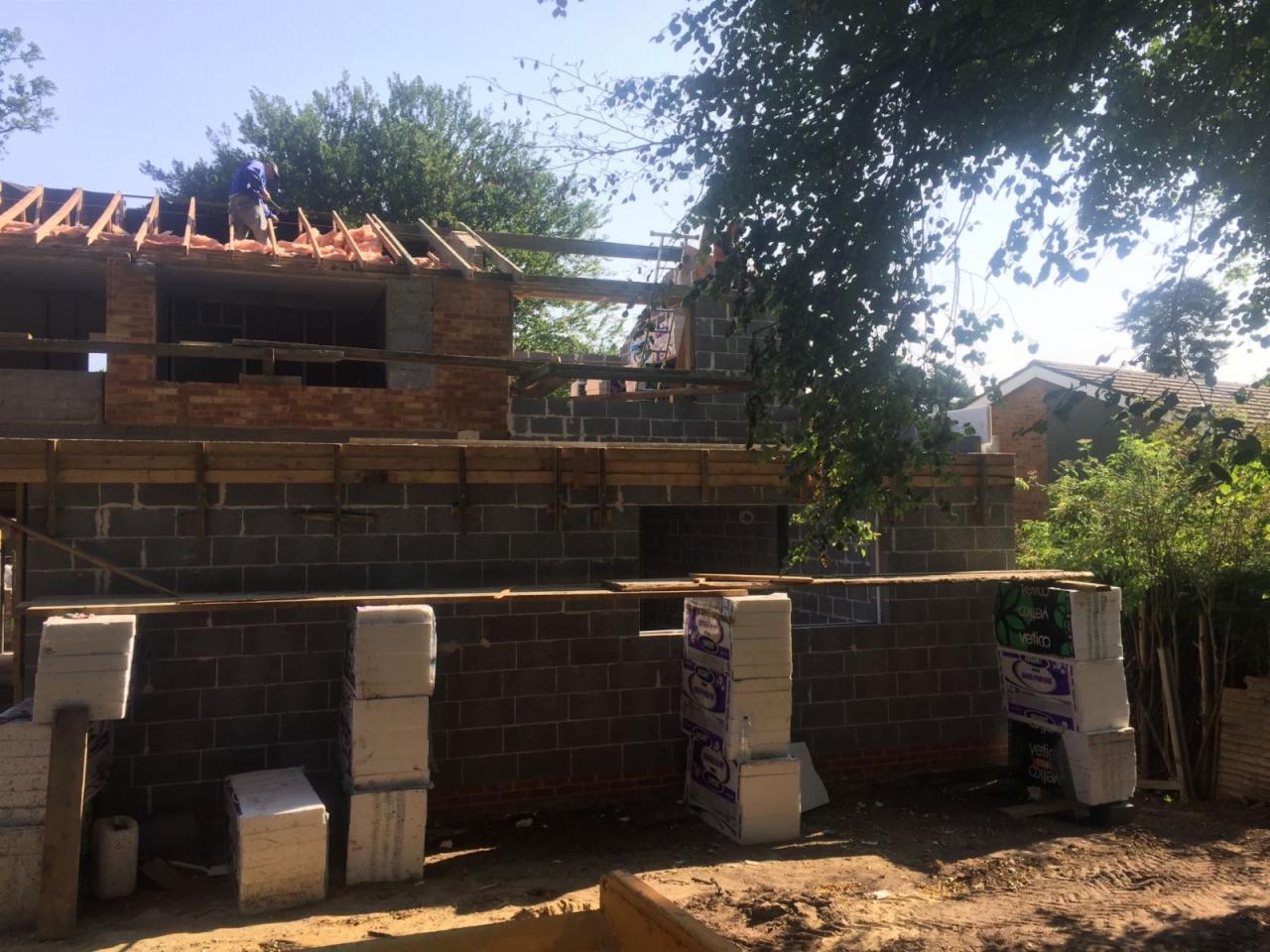 Rozbudowa domu w trakcie: murowane ściany z bloczków, drewniana konstrukcja dachu z ociepleniem, pracownik na dachu, materiały budowlane składowane obok ściany.