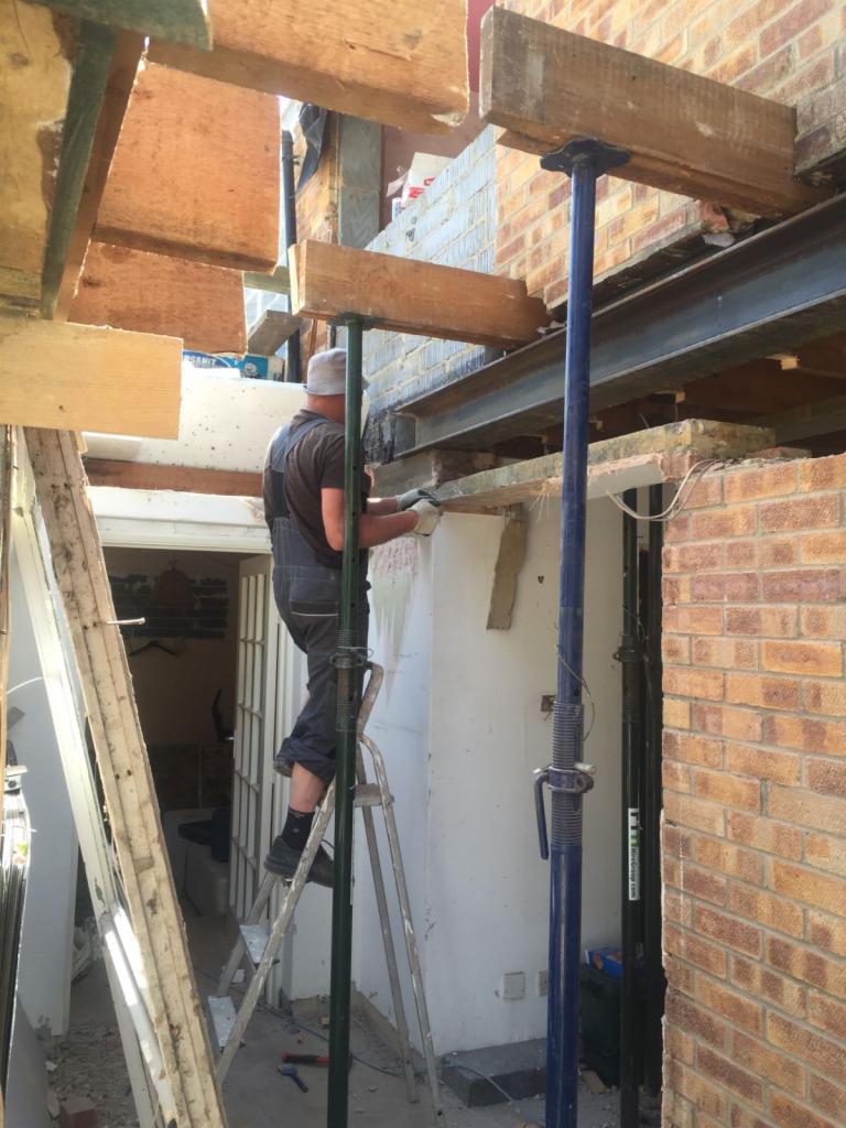 Pracownik na drabinie montuje drewniane podpory pod stalową belką podczas prac murarskich przy rozbudowie domu z cegły, widoczne narzędzia i wnętrze budynku.