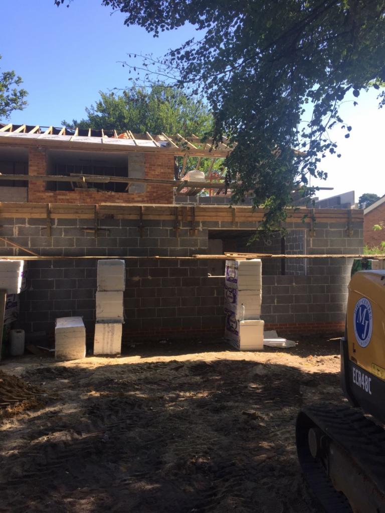 Wznoszenie murów parteru z bloczków betonowych, widoczne drewniane szalunki i częściowo zrealizowana konstrukcja dachu, na pierwszym planie stosy materiałów izolacyjnych i minikoparka.