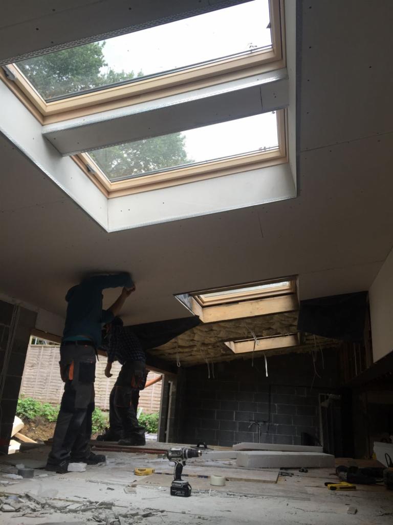 Montaż płyt gipsowo-kartonowych na suficie z oknami dachowymi, dwóch pracowników w odzieży roboczej, widoczny wkrętak akumulatorowy na podłodze, surowy stan wykończenia.