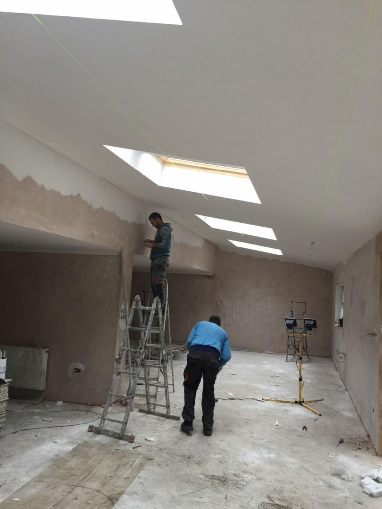Prace wykończeniowe wewnątrz domu z oknami dachowymi: dwóch pracowników na drabinie i podłodze, ściany w trakcie szpachlowania, widoczne narzędzia i oświetlenie budowlane.
