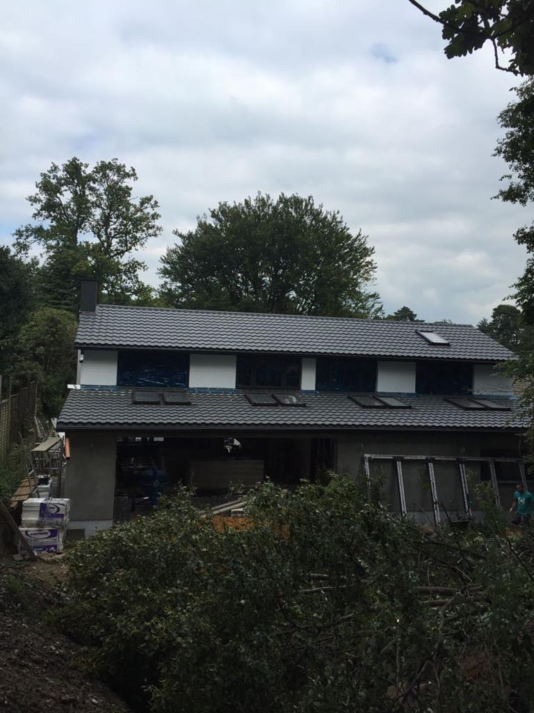 Dom w trakcie rozbudowy w Londynie, widoczna nowa część z szarą dachówką i oknami dachowymi, otoczony zielenią.