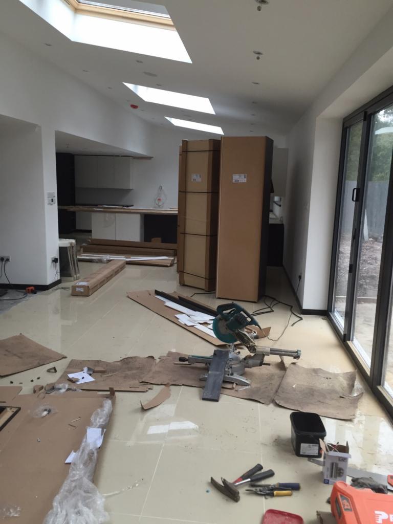 Wnętrze domu w trakcie prac wykończeniowych: widoczne kartony z wyposażeniem kuchennym, narzędzia do cięcia i montażu, oraz duże okno przesuwne.