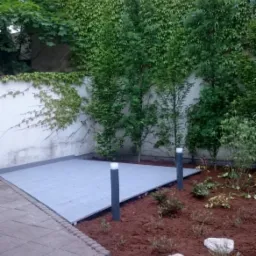 Nowoczesny taras z szarej deski kompozytowej, oświetlony niskimi lampami, otoczony świeżo nasadzoną roślinnością i korą, z murem porośniętym bluszczem w tle.