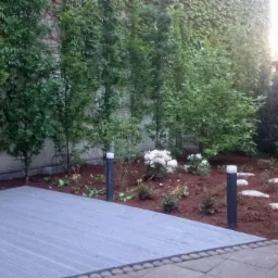 Nowoczesny ogród z drewnianym tarasem, oświetleniem punktowym, ścieżką z kamieni i ścianą porośniętą bluszczem.