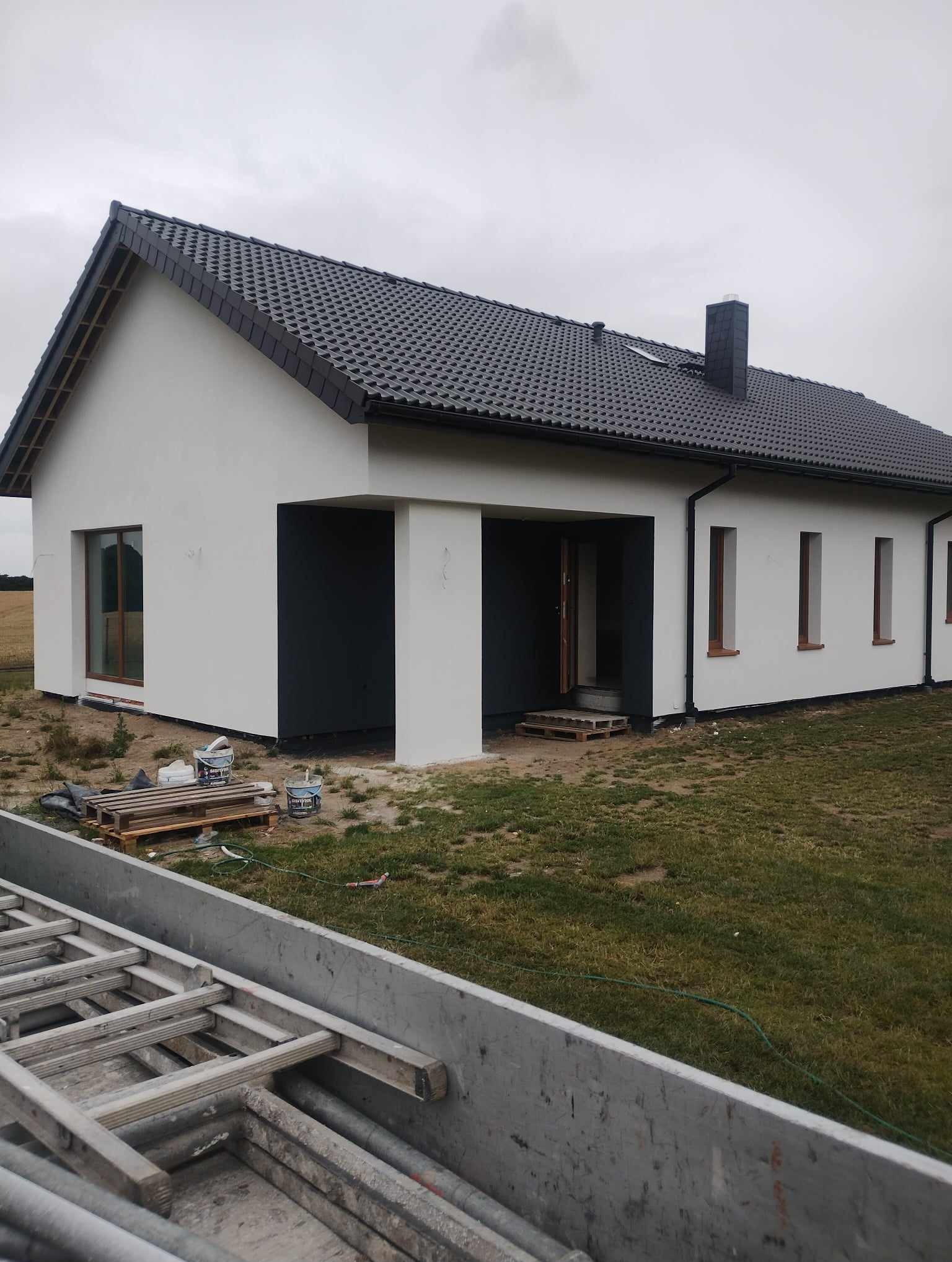 Nowo wybudowany dom jednorodzinny z szarą dachówką, świeżo pomalowany na biało z szarymi elementami, widoczne drabiny i puszki z farbą na palecie, trawa na posesji.