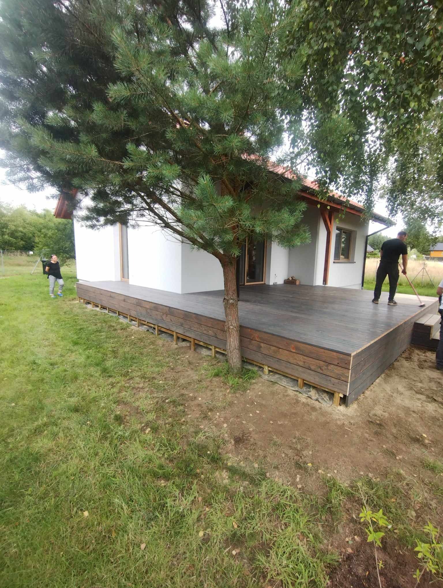 Nowo wybudowany, ciemnobrązowy taras drewniany przy białym domu jednorodzinnym, z widocznym podkonstrukcją i osobą zamiatającą powierzchnię.