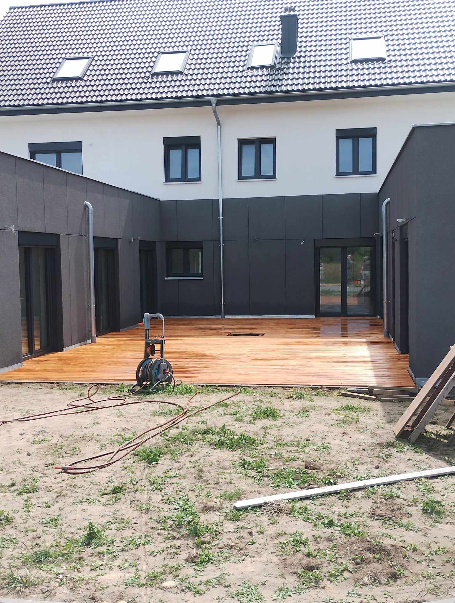 Świeżo położony, mokry drewniany taras przydomowy z widoczną myjką ciśnieniową na pierwszym planie, dom z szarą elewacją i czarnymi oknami w tle.