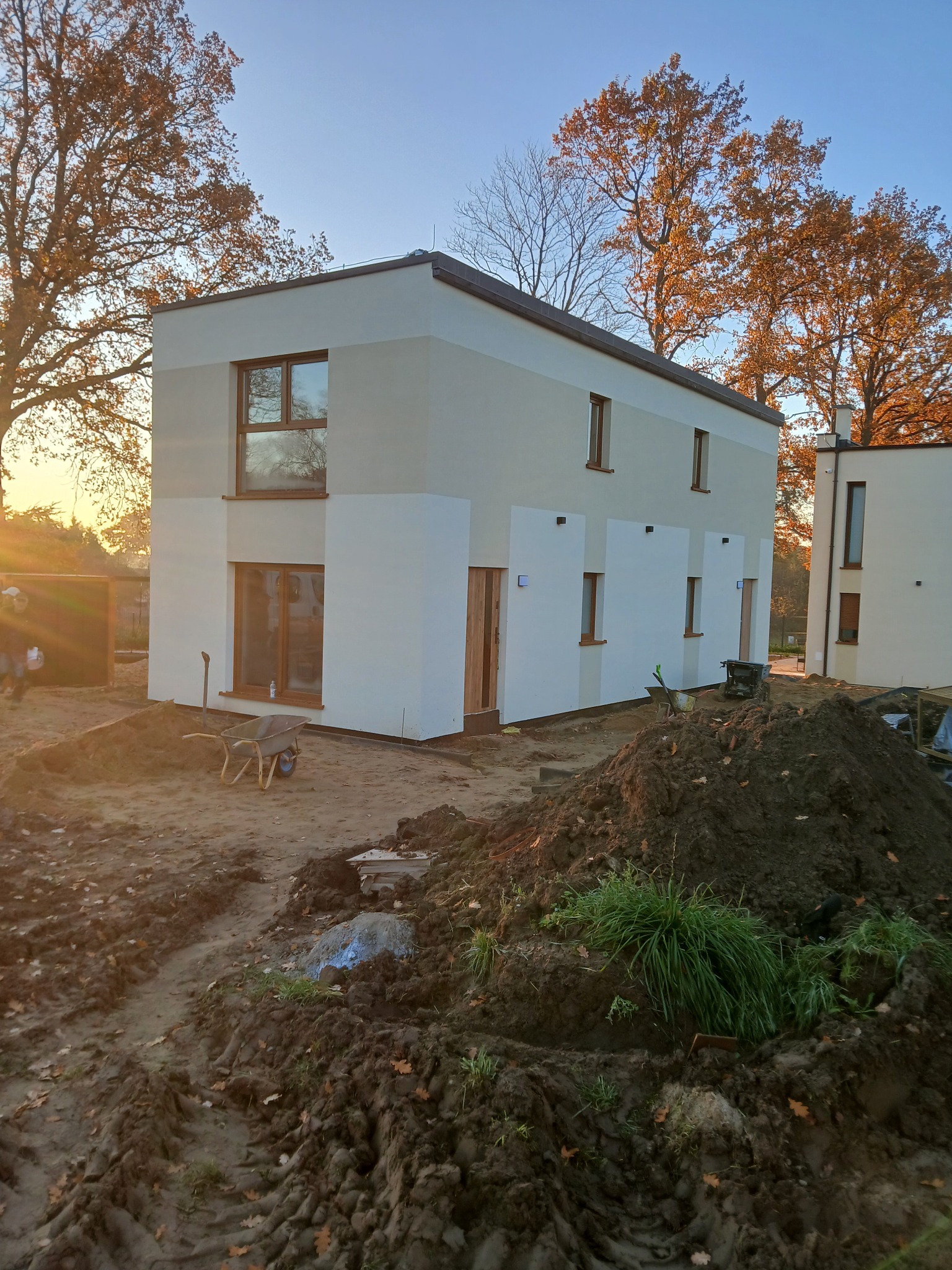 Nowoczesny, dwukondygnacyjny dom z jasną elewacją w trakcie budowy, widoczny w jesiennej scenerii z narzędziami budowlanymi i stertą ziemi na pierwszym planie.