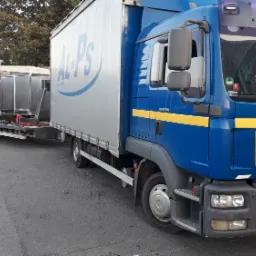 Niebieska ciężar&oacute;wka MAN z napisem 'POMOC DROGOWA' i przyczepą z metalowymi kontenerami na parkingu, widoczny numer rejestracyjny GWE 7919J.