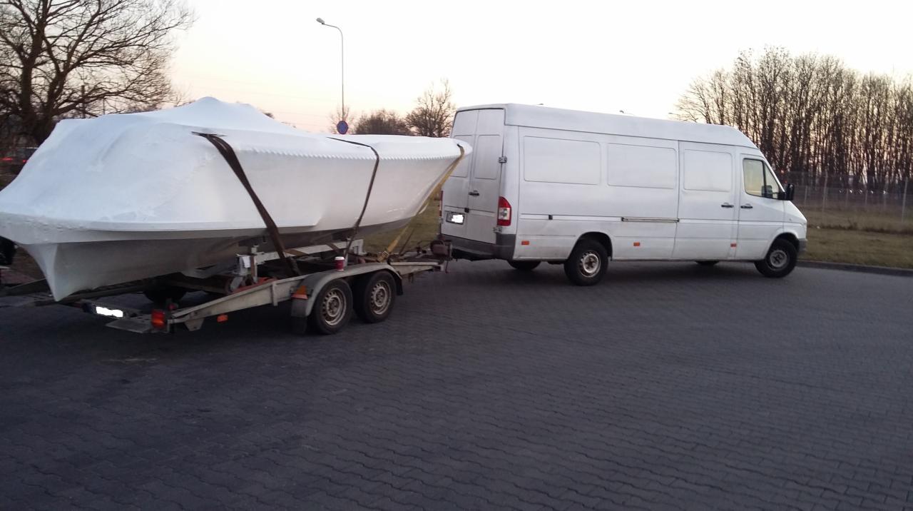 Biały van ciągnie przyczepę z łodzią pod plandeką, zaparkowany na szarym bruku, w tle drzewa i zachodzące słońce.