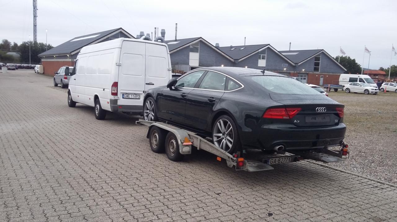 Czarny samochód Audi A7 na niskopodwoziowej przyczepie, holowany przez biały samochód dostawczy na tle budynków z szarymi dachami.