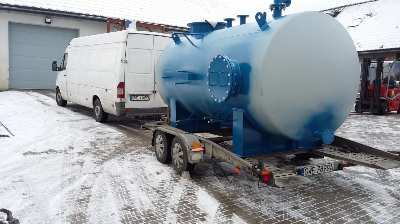 Biały bus z przyczepą przewożący duży, niebiesko-biały zbiornik w zimowej scenerii, z widocznym wózkiem widłowym w tle.